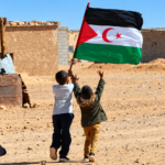 Two Sahrawi children carrying the Western Sahara flag in a desert settlement.