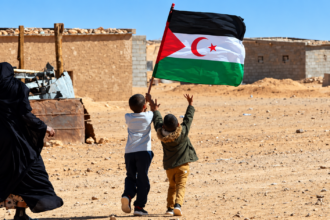 Two Sahrawi children carrying the Western Sahara flag in a desert settlement.
