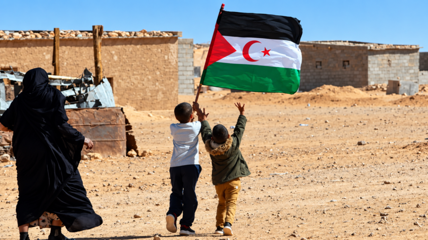 Two Sahrawi children carrying the Western Sahara flag in a desert settlement.